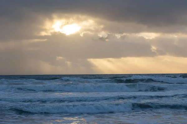 Foamy sea waves and sun peaking through the clouds at early evening. Mid shot