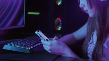 Cute gamer girl scrolling through her phone while sitting by PC. Mid shot