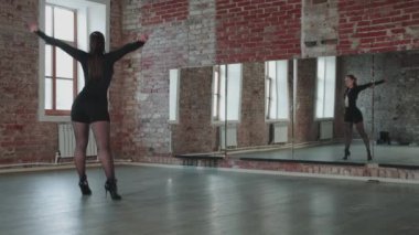 An attractive woman with nice body and wearing sexy black outfit practicing her dancing in front of a mirror in the studio. Mid shot