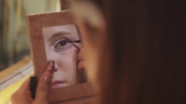 A cute girl doing makeup - drawing winged eyeliner looking in a small mirror. Mid shot
