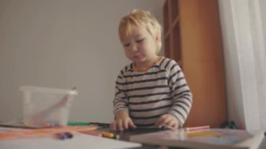 Cute little blonde boy playing with colored pencils by the table. Mid shot
