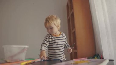 Cute little blonde boy collecting colored pencils from the table. Mid shot