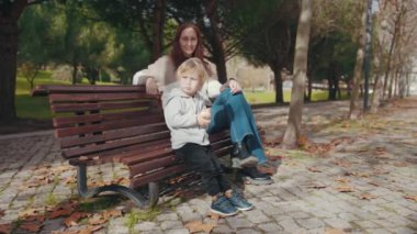 Woman with her little son in the park - sitting on the bench and the boy eating an apple. Mid shot