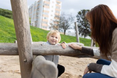 A little funny boy with his mother playing on the playground with wooden logs. Mid shot