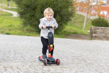 A little blonde boy riding a scooter in the park. Mid shot