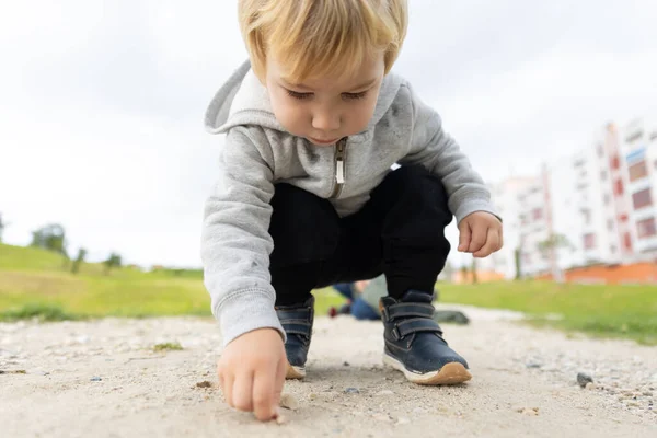 A little blonde boy bending down to the ground to pick up a little pebble. Mid shot