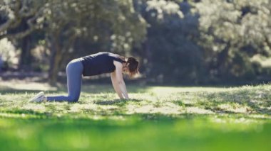 Spor kadını doğada yoga yapıyor. Sırtını esnetiyor ve çimlere uzanıyor. Orta çekim