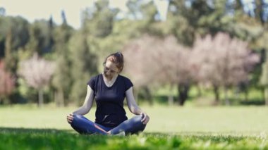 Doğa üzerine yoga yapan gözlüklü bir kadın lotus pozisyonunda oturup boynunu ısıtıyor. Orta çekim
