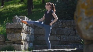 Atletik kadın ayağını taş bir duvara atarak esneme hareketleri yapıyor. Orta çekim