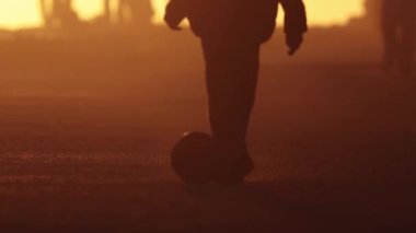 Gün batımında siluetleri olan bir çocuk topa vuruyor. Orta çekim