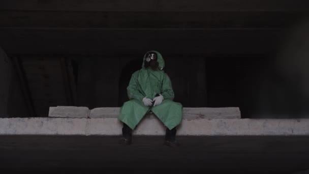 启示录后的概念 一个戴着防毒面具和防护服的男人坐在混凝土板上摆动着他的腿 — 图库视频影像
