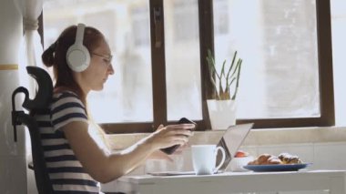 Kulaklık takan, bilgisayarını ve telefonunu kullanarak mutfakta çalışan yetişkin bir iş kadını. Orta çekim