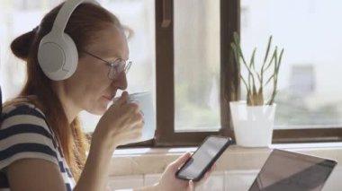 Kulaklıklı bir iş kadını mutfakta çalışıyor, telefonunu kullanıyor ve kahve içiyor. Orta çekim