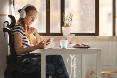 Kulaklıklı yetişkin bir iş kadını mutfak masasında oturup telefonuna bakıyor. Orta çekim