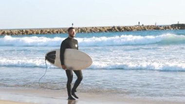 Büyük bir sörf tahtasıyla kıyı boyunca yürüyen gülümseyen adam. Orta çekim