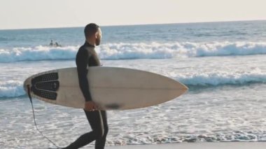 Büyük bir sörf tahtasıyla sahil boyunca yürüyen bir adam. Orta çekim