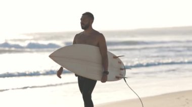 Çıplak gövdeli çekici bir adam deniz kıyısında sörf tahtasıyla yürüyor. Orta çekim