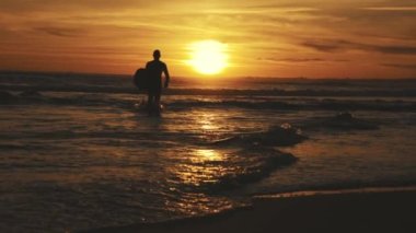 Gün batımında sörf tahtasıyla denize açılan atletik bir adamın silueti. Orta çekim