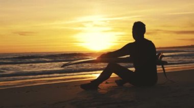 Gün batımında deniz kıyısında oturan bir sörfçünün silueti. Orta çekim