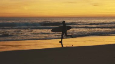 Gün batımında sörf tahtasıyla deniz kıyısında ağır çekimde koşan atletik bir adam. Orta çekim
