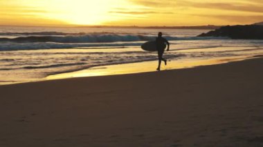 Gün batımında sörf tahtasıyla deniz kıyısında koşan atletik bir adam. Orta çekim