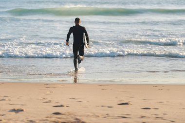 Dalgıç kıyafetli bir sporcu elinde sörf tahtasıyla okyanusa koşuyor. Orta çekim