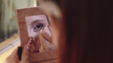 Genç bir kadın paletindeki aynaya bakarak kanatlı göz kalemi çiziyor. Orta çekim