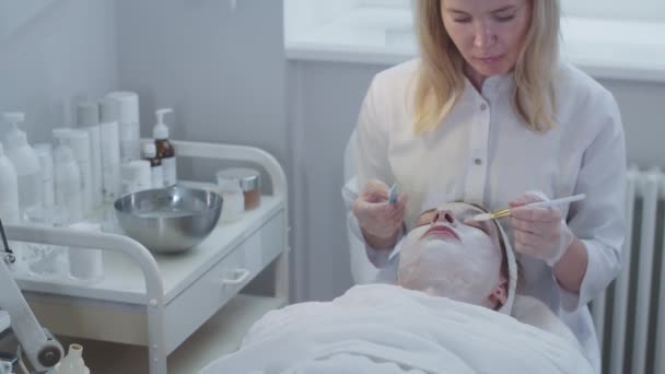Esteticista Aplica Máscara Branca Rosto Seu Cliente Usando Pincel Meio — Vídeo de Stock