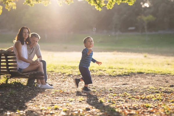公园里可爱的白人家庭 一个追逐肥皂泡的小男孩和他的父母在看着他 — 图库照片