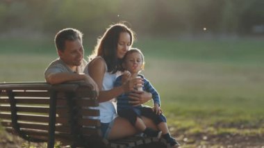 Ebeveynler bir bankta oturup oğullarını sabun köpüğüyle oynuyorlar. Orta çekim