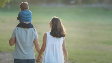 Parkta yürüyen sevimli genç bir aile. Babasının omuzlarında küçük bir çocuk. Orta çekim