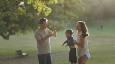 Şirin, genç bir aile parkta vakit geçiriyor. Baba sabun köpüğü üflüyor ve anne oğlunu havada büyütüyor. Orta çekim