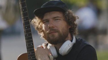 Elinde gitar tutan kablosuz kulaklık takan bir adam kameraya bakıyor. Portre