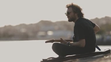 Rıhtımda gitar çalan bir erkek müzisyen. Orta çekim