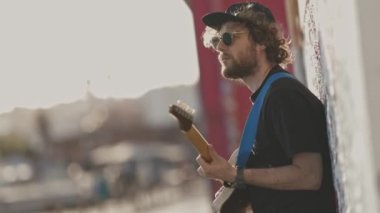Duvarın yanında durup gitar çalan bir adam. Orta çekim
