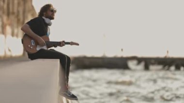 Rıhtımda oturup gitar çalan bir adam. Orta çekim