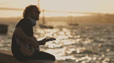 Kablosuz kulaklık takan bir adam gün batımında rıhtımda oturup gitar çalıyor. Orta çekim