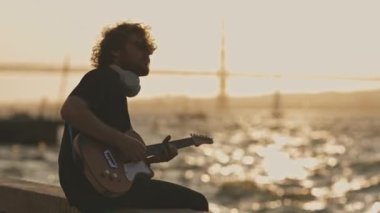 Rıhtımda oturan ve günbatımında elektro gitar çalan kablosuz kulaklık takan bir adam. Orta çekim