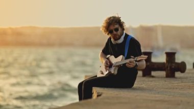 Bir adam, bacakları suya sarkık bir şekilde sette oturur ve gitar çalar. Orta çekim