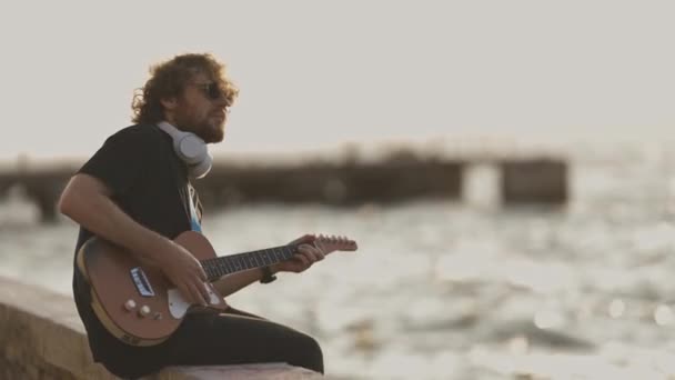 一个戴着无线耳机坐在海滨弹吉他的男人 — 图库视频影像