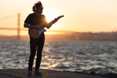 Bir adam rıhtımda duruyor ve parlak turuncu gün batımında gitar çalıyor. Orta çekim