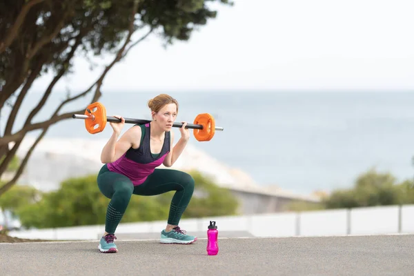 stock image Adult athletic woman squatting with a dumbbell on her shoulders. Mid shot