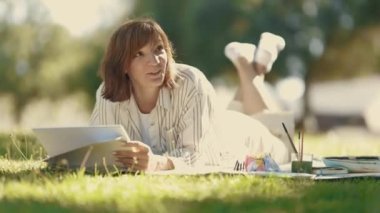 Parktaki battaniyeye uzanmış elinde çizim defteriyle birileriyle konuşan yetişkin bir kadın. Orta çekim