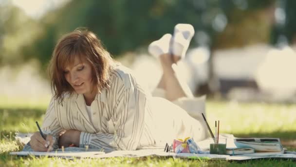 Взрослая Улыбающаяся Женщина Лежит Одеяле Парке Рисует Альбоме Средний Выстрел — стоковое видео
