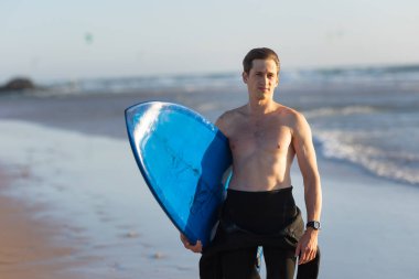 Deniz kıyısında elinde sörf tahtası olan çıplak vücutlu zinde bir adam. Orta çekim