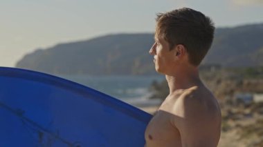 Plajda çıplak gövdeli bir adam sörf tahtası tutuyor. Portre