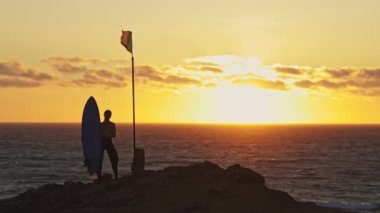 Okyanus ve günbatımının arka planında sörfçü bir adamın silueti. Rüzgarda dalgalanan Portekiz bayrağı. Orta çekim