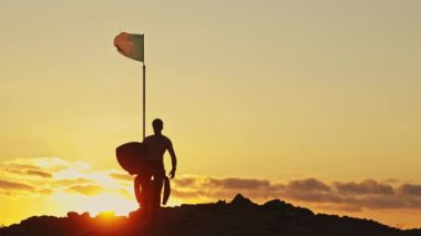 Gün batımında kayada yürüyen bir sörfçünün silueti. Rüzgarda sallanan Portekiz bayrağı. Orta çekim