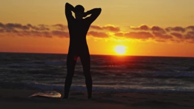 Bir adam dalgıç kıyafetini bağlar ve kumdan sörf tahtasını kaldırır. Parlak günbatımı. Orta çekim