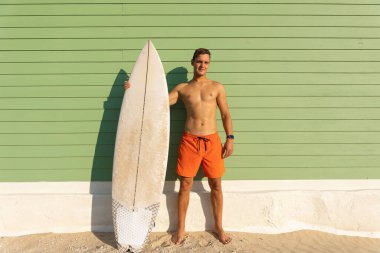 Açık yeşil duvarda elinde sörf tahtası olan hoş vücutlu bir adam. Orta çekim